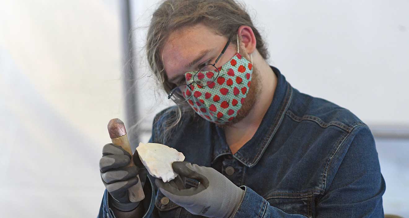 Beth Cooper ’24 demonstrates the craft of flintknapping during a “Pumpkin Carving with Stone Tools” outreach program. The self-taught knapper developed an interest in the craft their senior year of high school to “try something new.” Cooper had already had dabbled in blacksmithing, metal casting, and woodworking. “While I enjoy doing all of those, they can be difficult skills to maintain without a workshop or money for supplies and tools, so I started teaching myself to knap. It’s cheaper and less seasonal than my other hobbies,” they said. Pictured, Cooper is using a “copper bopper” to knap a piece of Keokuk chert from a flintknapping supplier and quarry in Oklahoma.