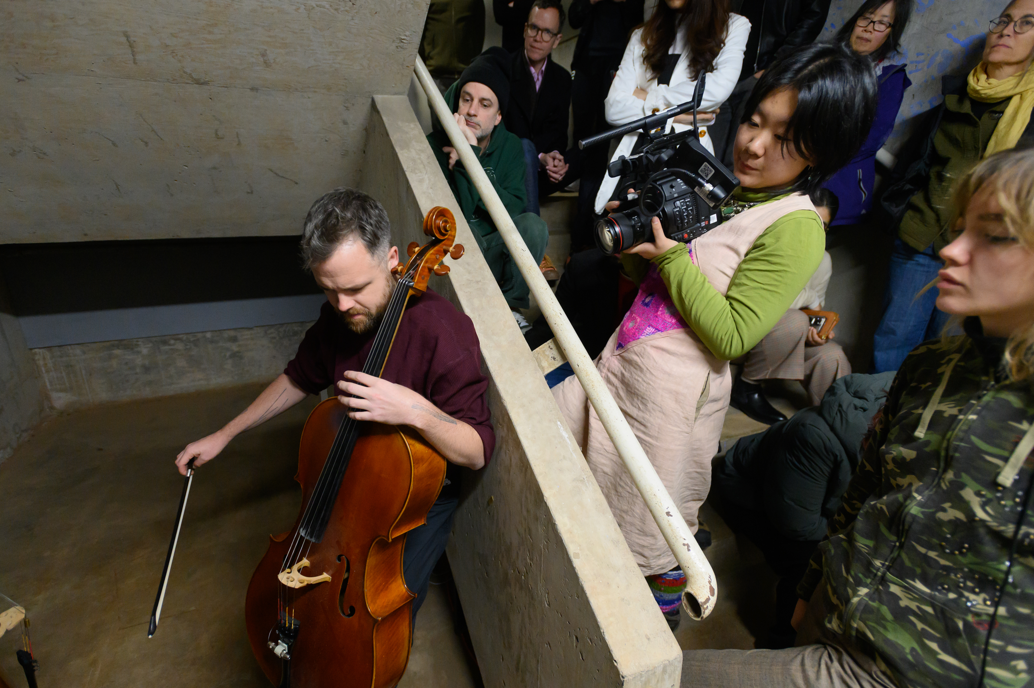A man plays a cello in a cramped tunnel while an audience watchs