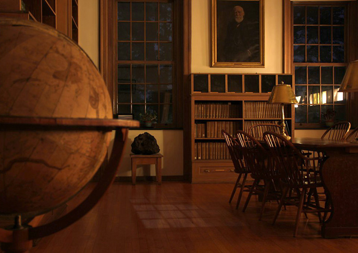 The warmly lit observatory library with wooden shelves, a large globe, and tall windows.