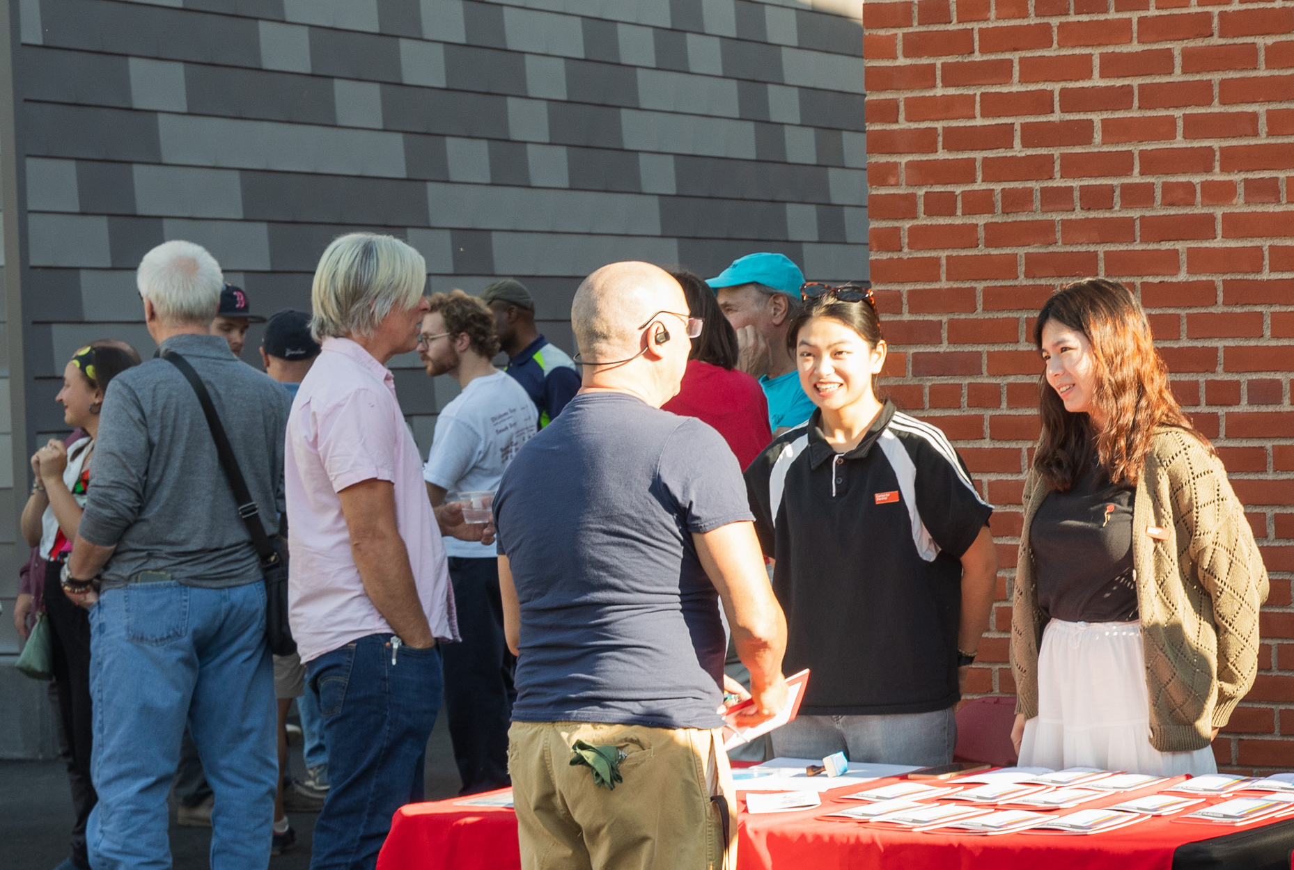 Two students behind table with handouts talk to man seen from behind with other people standing and talking in the background 