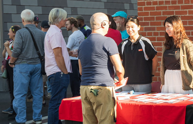 Two students behind table with handouts talk to man seen from behind with other people standing and talking in the background 