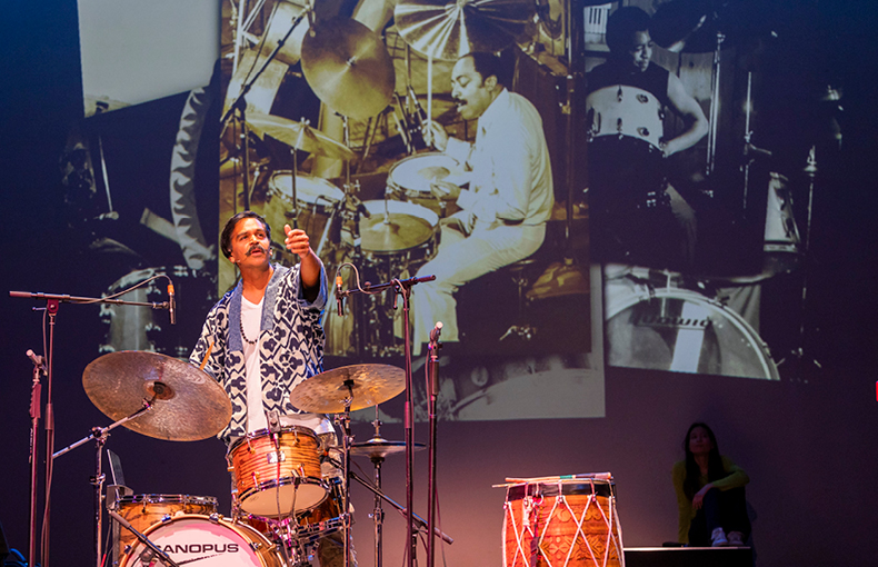 Musician on stage with drumset and dhol with images of jazz drummers displayed behind him