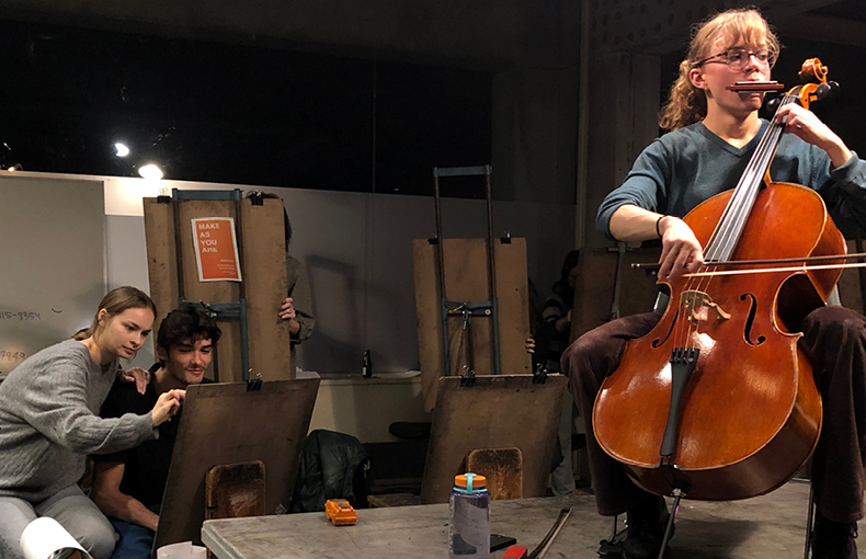 In drawing studio, two seated students draw while another student plays cello with harmonica while seated on a platform