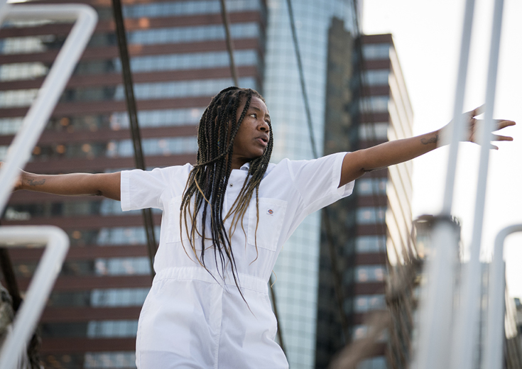 Dancer standing on sailboat with arms outstretched with rigging lines in front and buildings in background