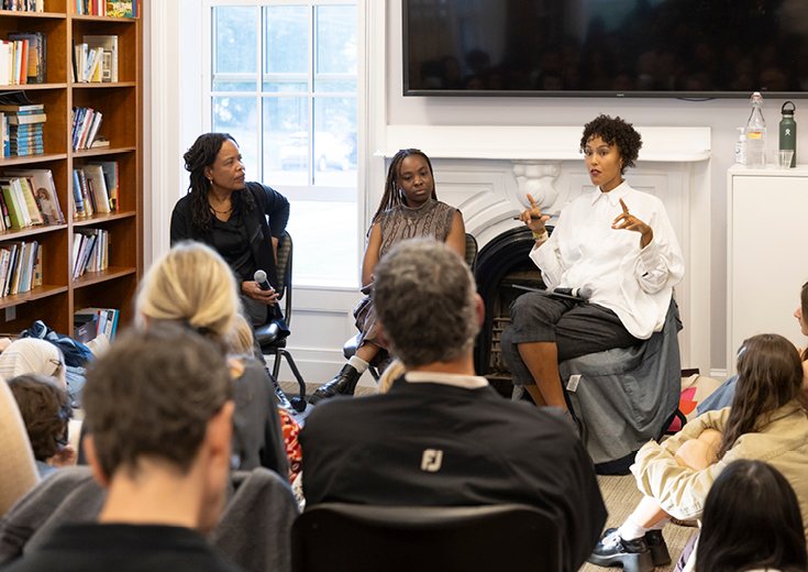 Three seated artists talk to audience in a room with bookshelves and fireplace and window in background
