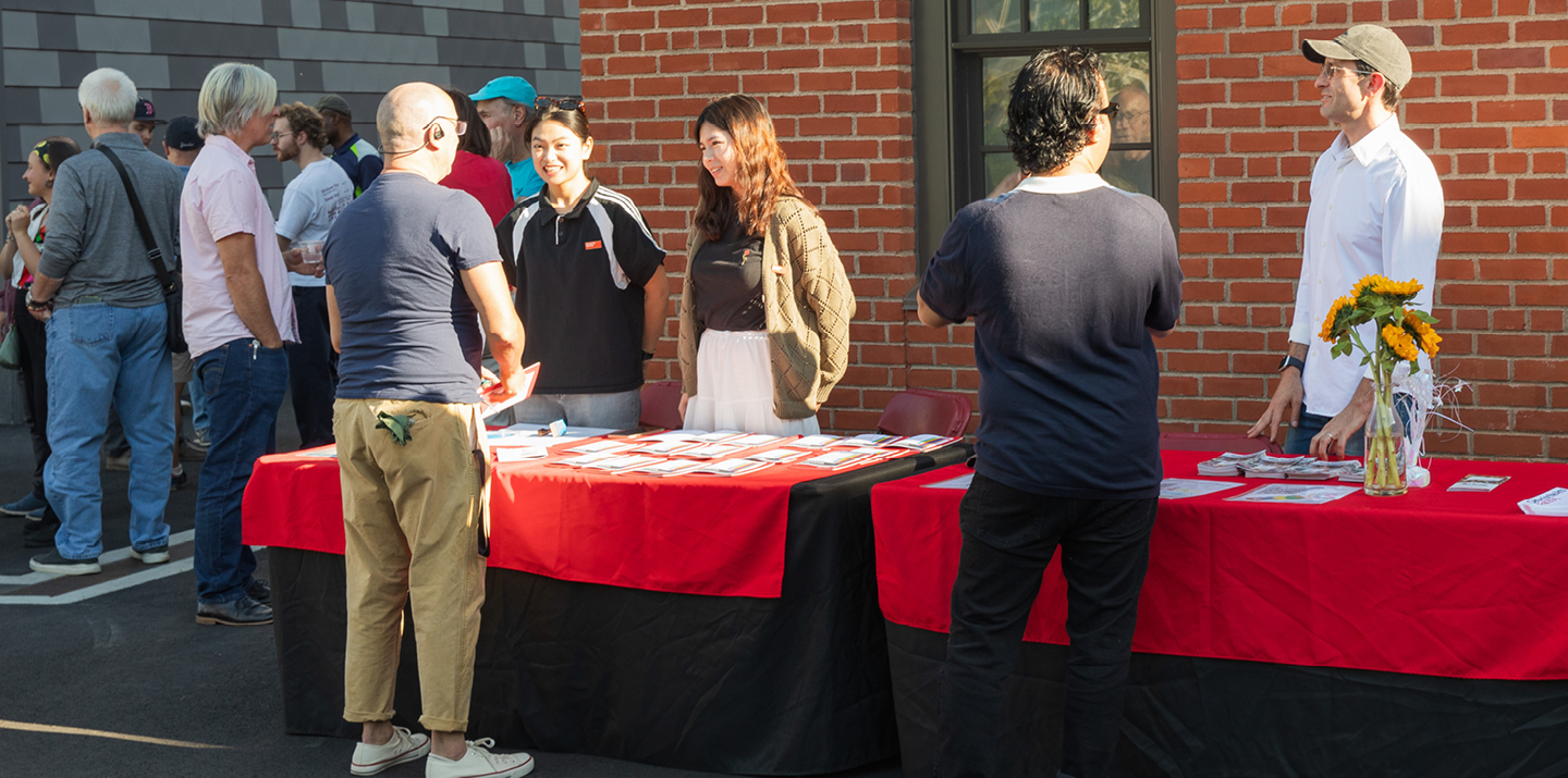 Two students behind table with handouts talk to man seen from behind with other people standing and talking in the background