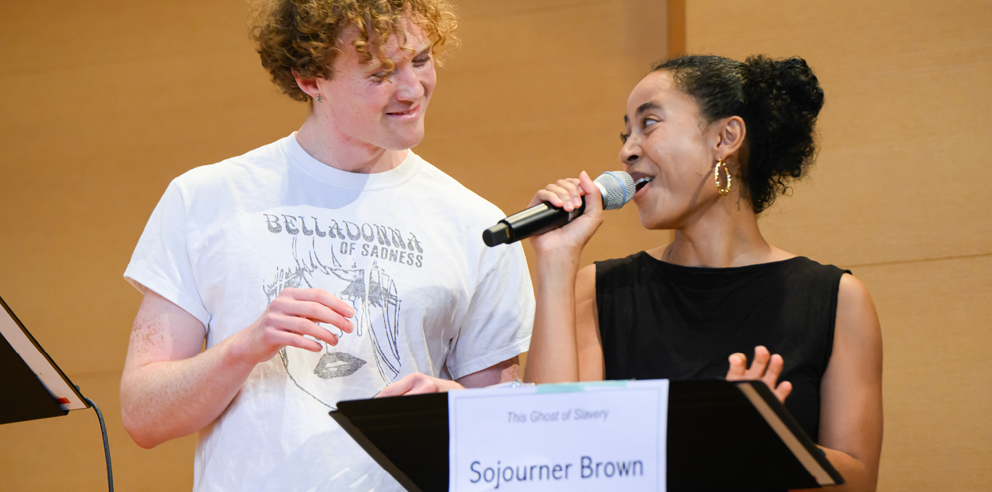 Two actors, one with microphone speaking, behind a music stand with a sign taped to the front that reads “The Ghost of Slavery” and “Sojourner Brown” 