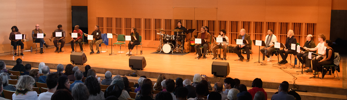 Actors seated on stage with music stands and a drummer in the center with audience watching from seats