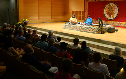 Three musicians sitting on rug on stage with tapestry depicting Saraswati behind them and audience in front