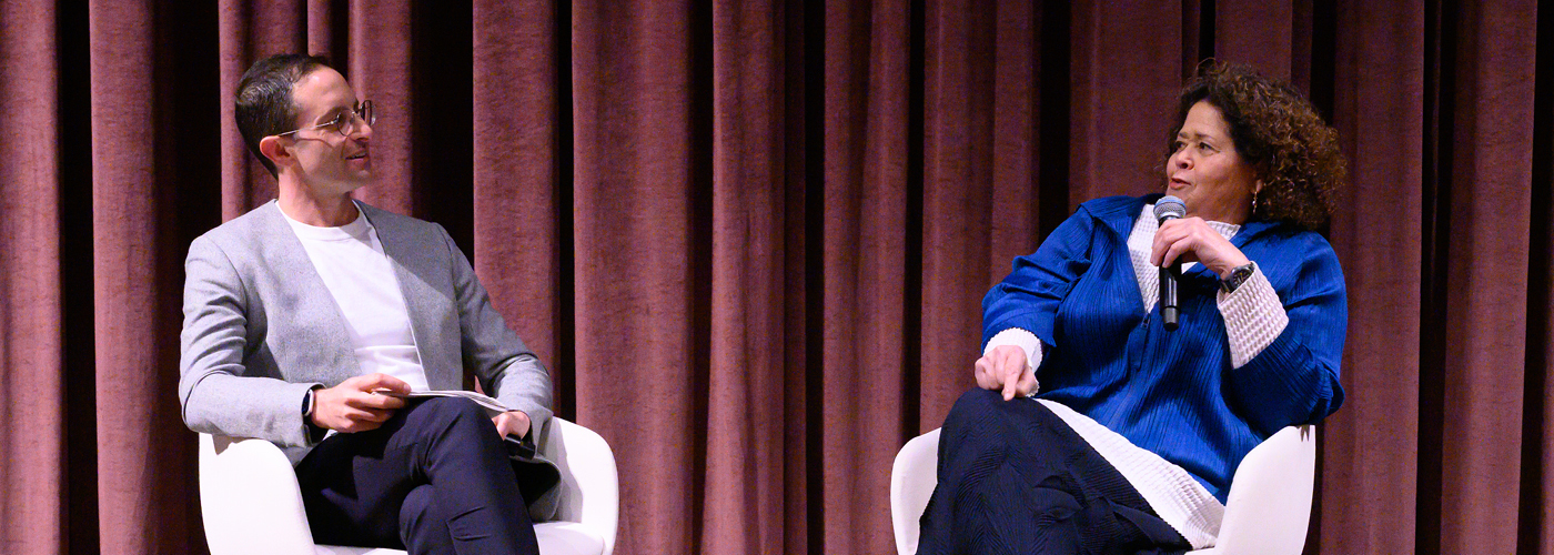 Two people with microphones seated in white chairs on stage 