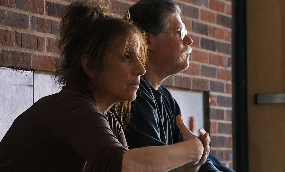 Two people seated with a brick wall behind