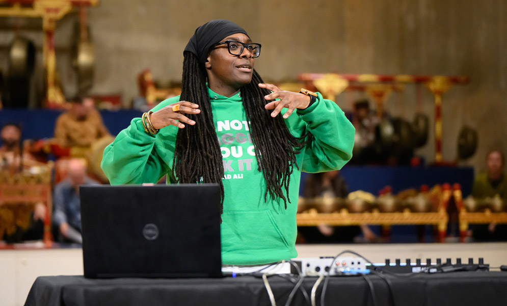 Artist with electronic instrument speaking to audience with Javanese gamelan in background