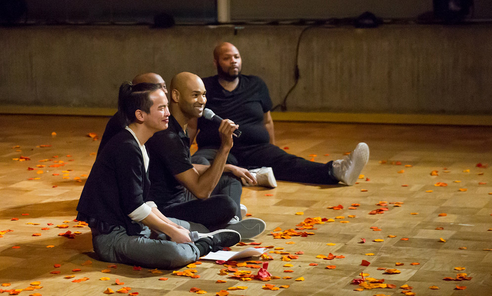 Artist holding microphone seated on floor with three others and flower petals