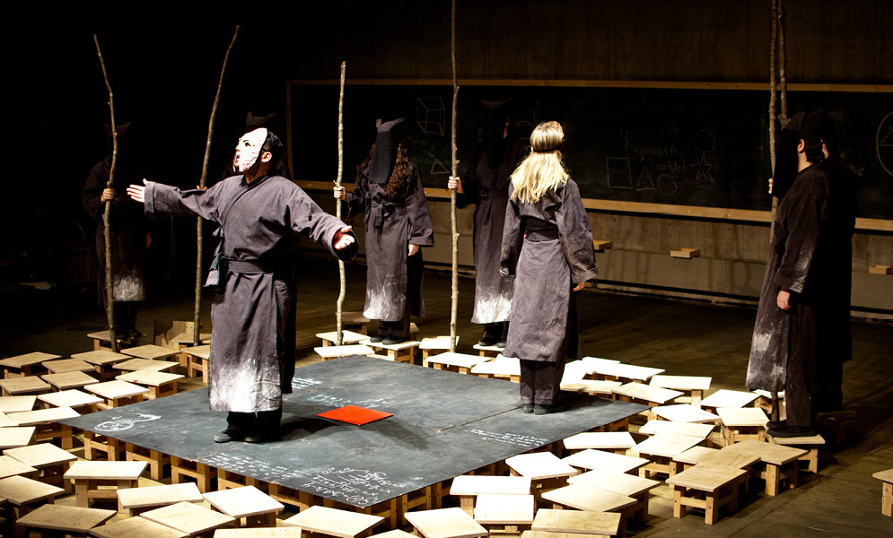 Actors with masks on and around platform, some holding sticks