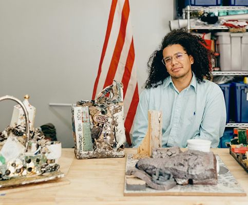 Artist seated at table with clay and sculptures on top and a shelf of materials in background