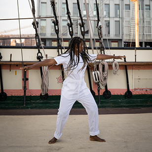 Dancer with arms outstretched on deck of sailboat with rigging lines seen behind and buildings in the distance