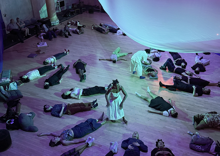 People lying on floor with dancer moving among them under suspended white sheet 