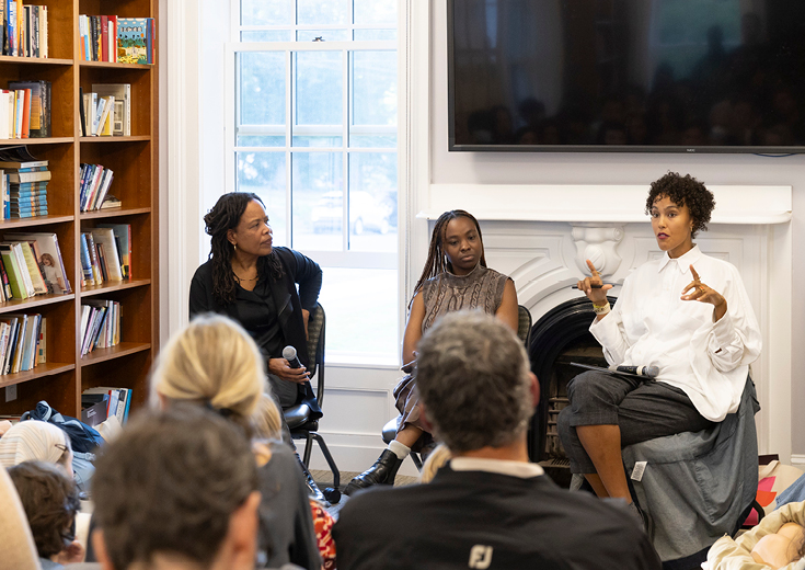 Three artists sitting in front of an audience seen from behind in a room with windows, fireplace, television screen, and bookshelves