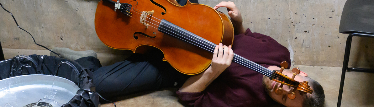Musician lying on cement floor with cello next to chair and fountain 