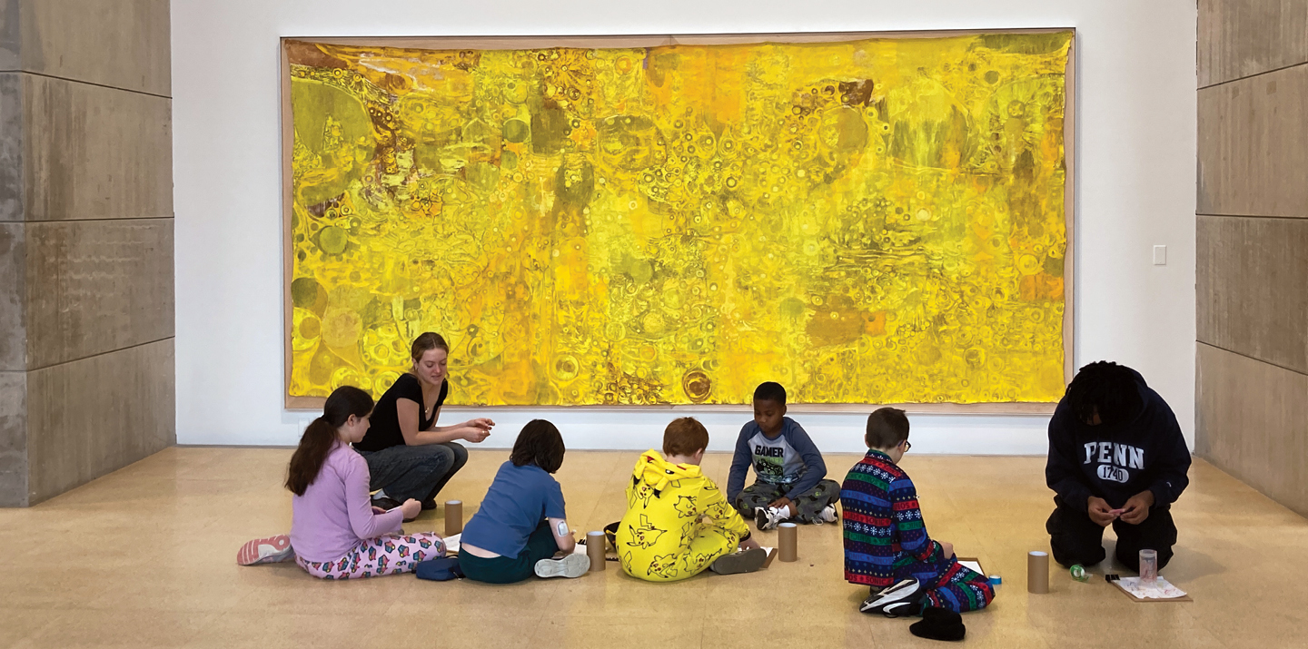 Student leads activity with younger students on gallery floor in front of large painting on wall