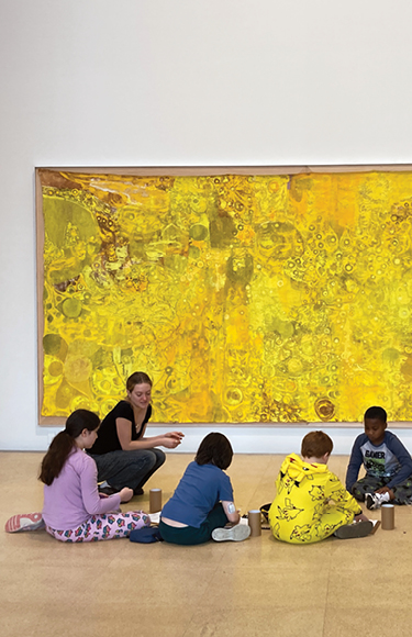 Student leads activity with younger students on gallery floor in front of large painting on wall - mobile version