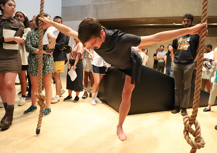 Dancer in gallery holds two ropes suspended from above while audience watches from all around