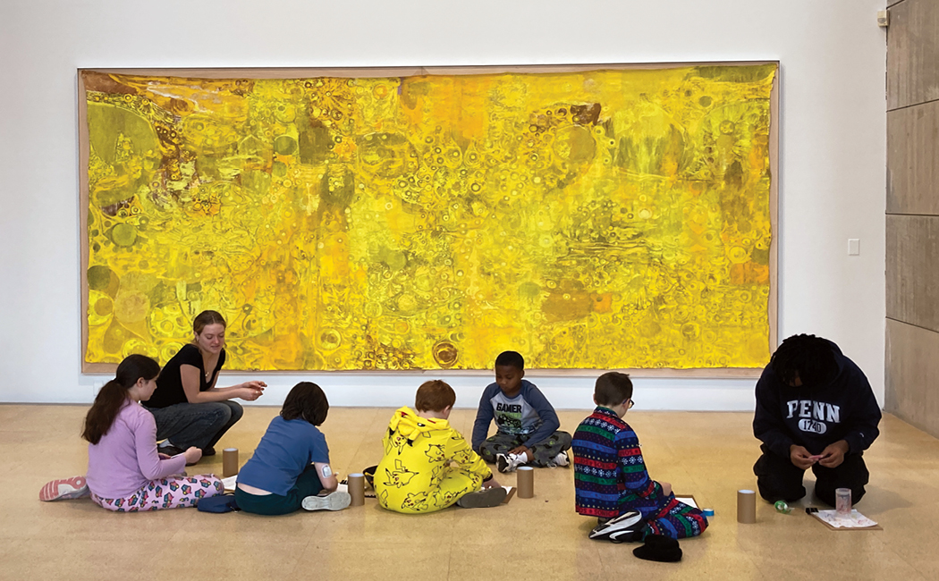 Student leads activity with six younger students on gallery floor in front of large painting on wall 