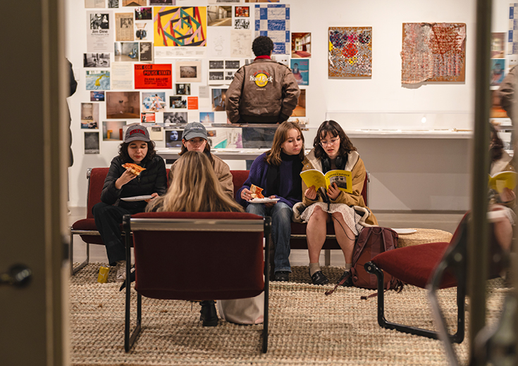 Students sitting on couch eating pizza and others behind looking at artwork on wall 