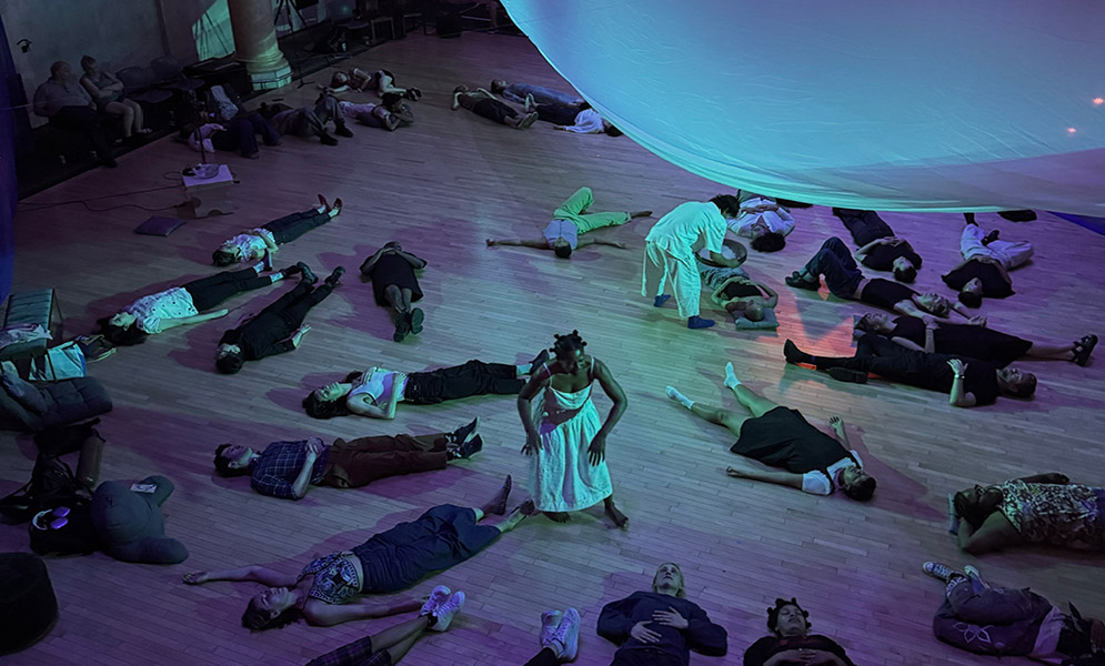 People lying on floor with two others walking between them with curved white cloth above