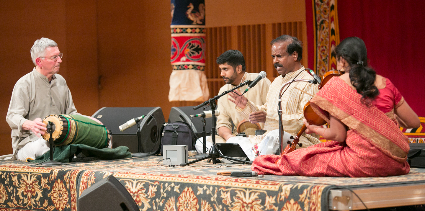 A singer, drummer, violinist, and percussionist perform while seated on a concert hall stage. 