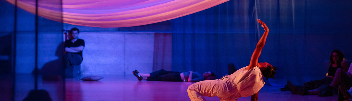  Dancer on floor reaching toward sheet suspended above with other people sitting and lying down in background 