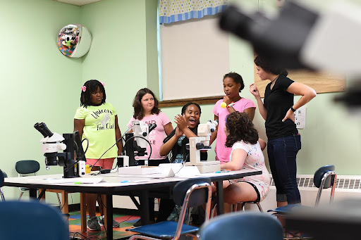 Wesleyan student explaining scientific concepts to young girls standing in front of microscopes. 