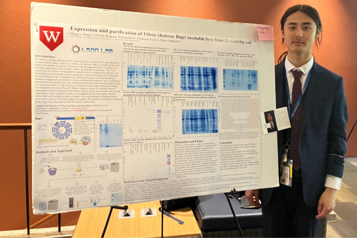 a young man in a suit stands beside his scientific poster